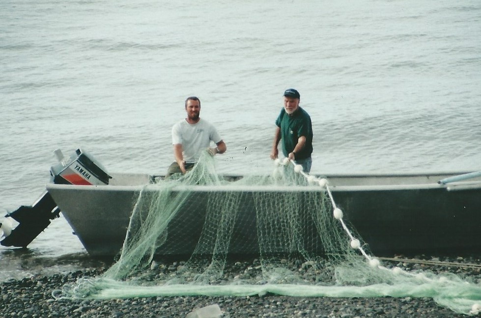 Two fishermen pulling a fishing net from a small boat on a rocky shoreline, with calm water in the background. The boat features a Yamaha motor, and the fishermen are engaged in their fishing activity.