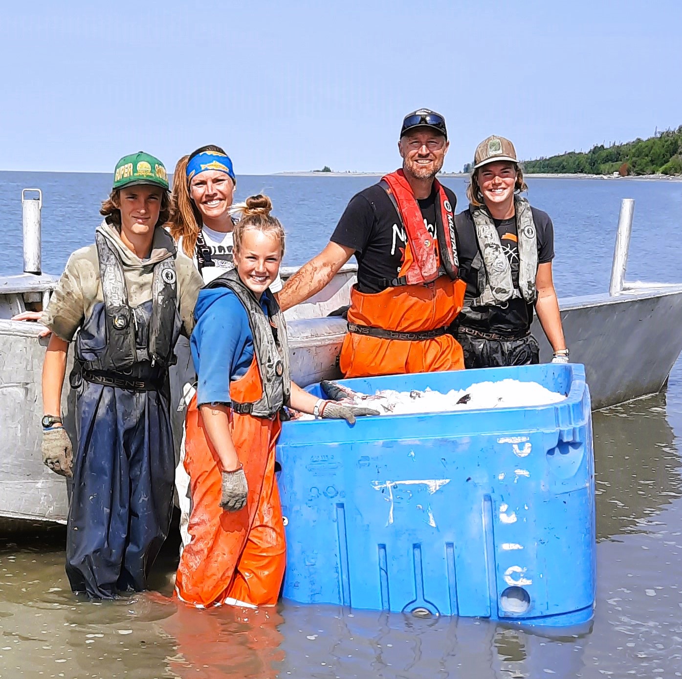Group of four individuals in fishing gear standing in shallow water beside a boat, smiling while posing with a blue container filled with fish. The scene captures a sunny day on the water, highlighting a family or team engaged in fishing activities.